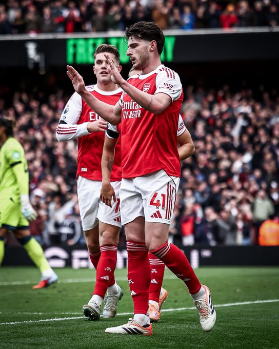 Two Arsenal players in red and white jerseys on a soccer field, waving to a crowd. One player wears the number 41 jersey. A goalkeeper in a green uniform is visible in the background, and a packed stadium with spectators is seen behind them.