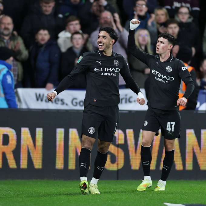 لاعبان يرتديان قمصان مانشستر سيتي السوداء مع شعار إيتهاد يحتفلان برفع أيديهما في فرح على ملعب عشبي أخضر مع جمهور في المدرجات الخلفية ولافتة بيضاء مكتوب عليها weses وأضواء برتقالية في الأسفل